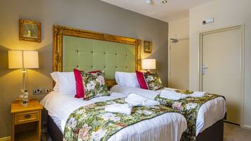Una habitación de hotel con dos camas, ropa de cama floral, un cabecero tapizado en verde y lámparas de noche.
