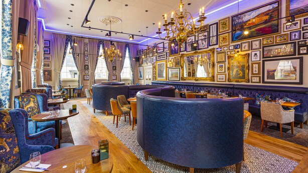 Restaurante com mesas redondas, assentos estofados em azul e parede decorada com quadros emoldurados.