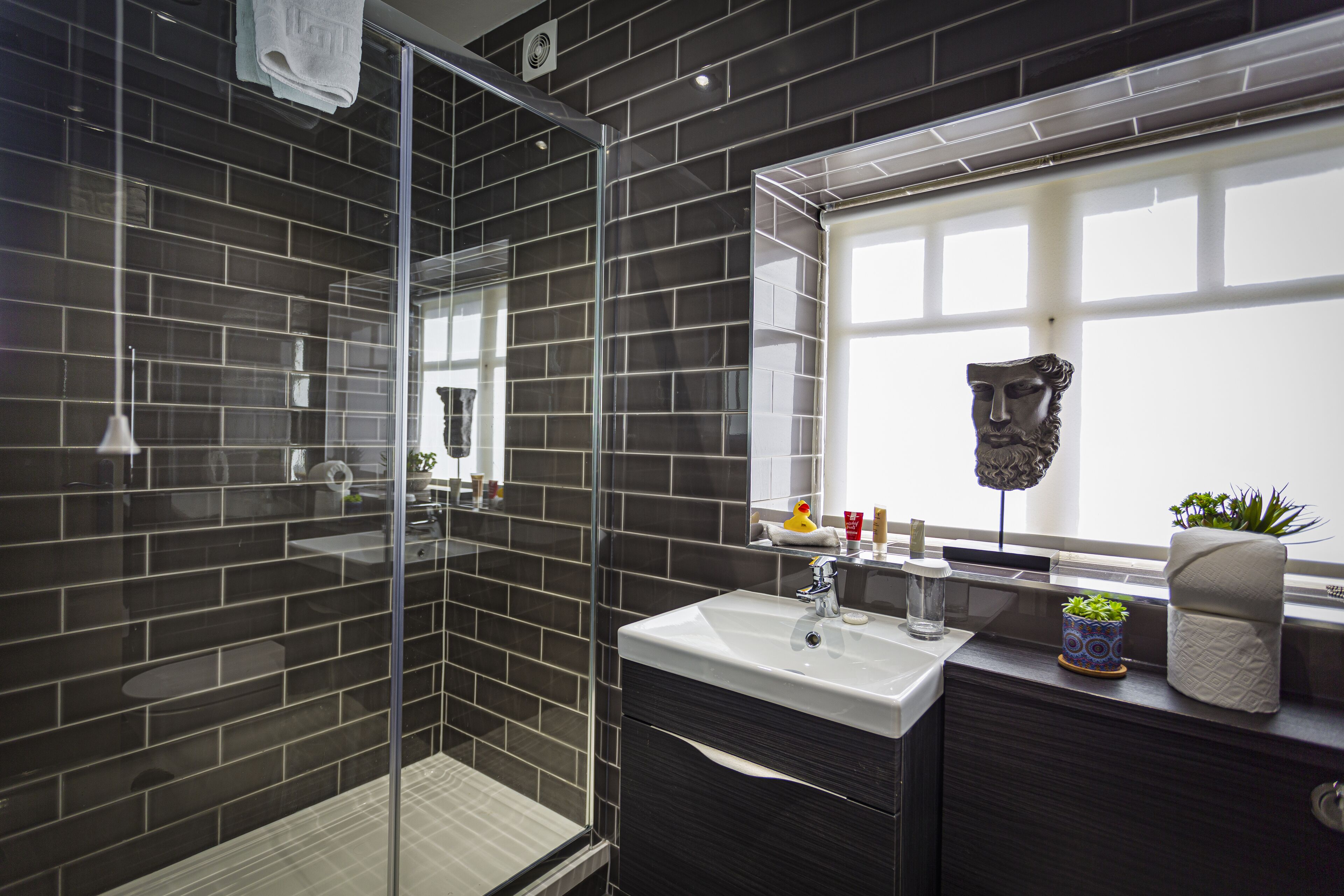 Un baño moderno con paredes de azulejos oscuros, cabina de ducha de vidrio, lavamanos blanco y ventana con vistas.