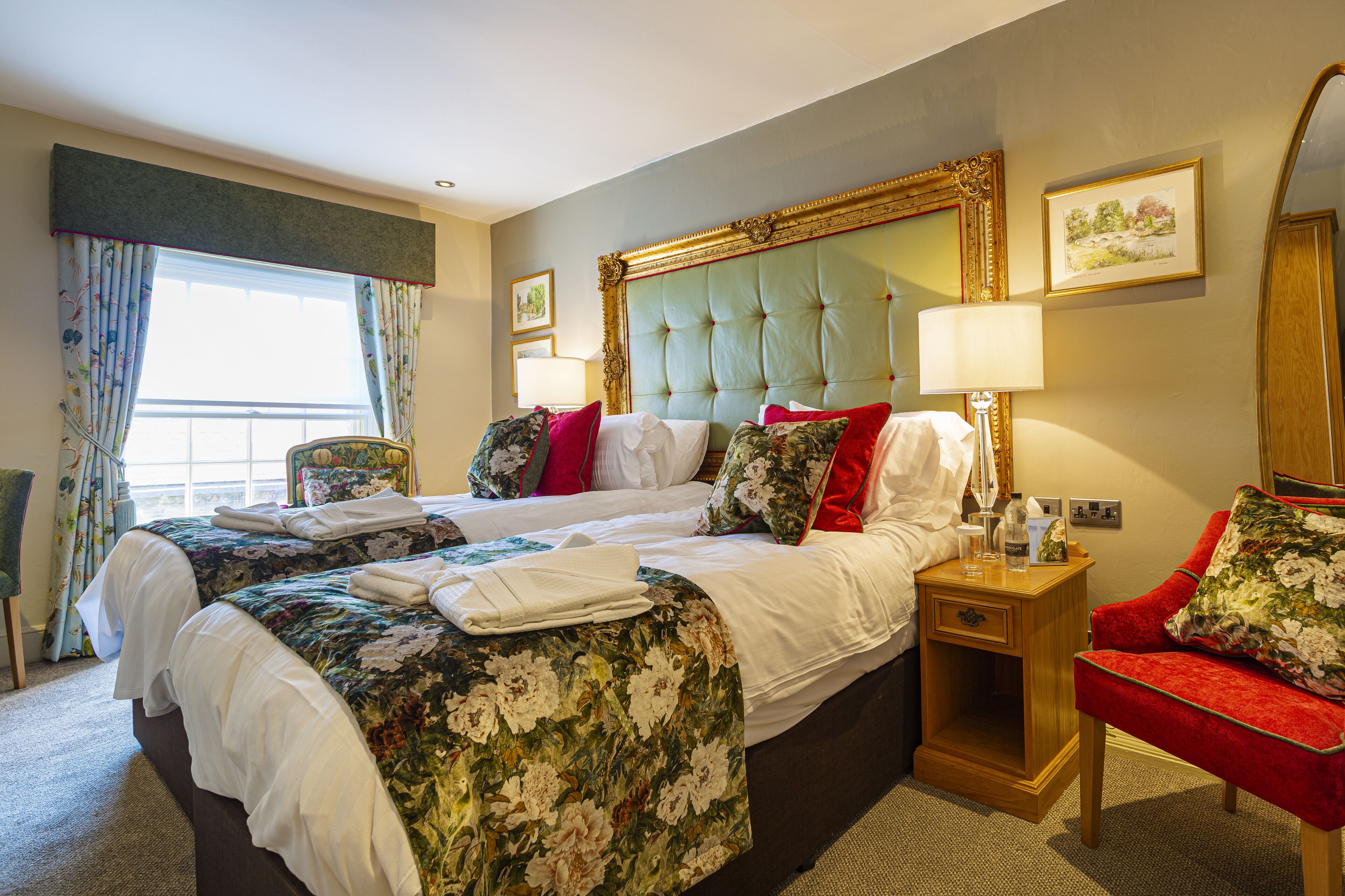 Habitación de hotel con una cama grande, un sillón rojo, una mesita de noche de madera y un ventanal con cortinas.