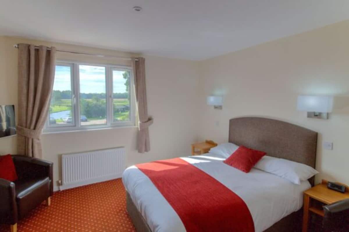 A hotel room with a bed, a chair, a desk, a window with curtains, and a view of the outdoors.