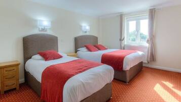 Habitación de hotel con dos camas, cada una con una manta roja, una mesita de noche de madera y una ventana con cortinas.