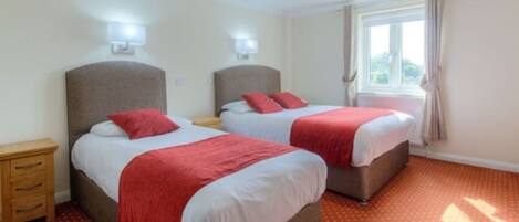 A hotel room with two beds, each with a red blanket, a wooden nightstand, and a window with curtains.