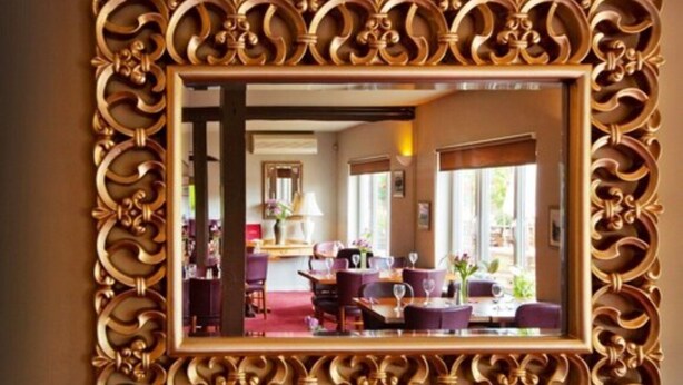 A mirror with an ornate gold frame reflecting a dining area with purple chairs and a table.