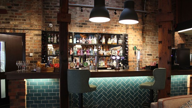 A bar area with a brick wall, shelves of liquor bottles, two bar stools, and a counter.