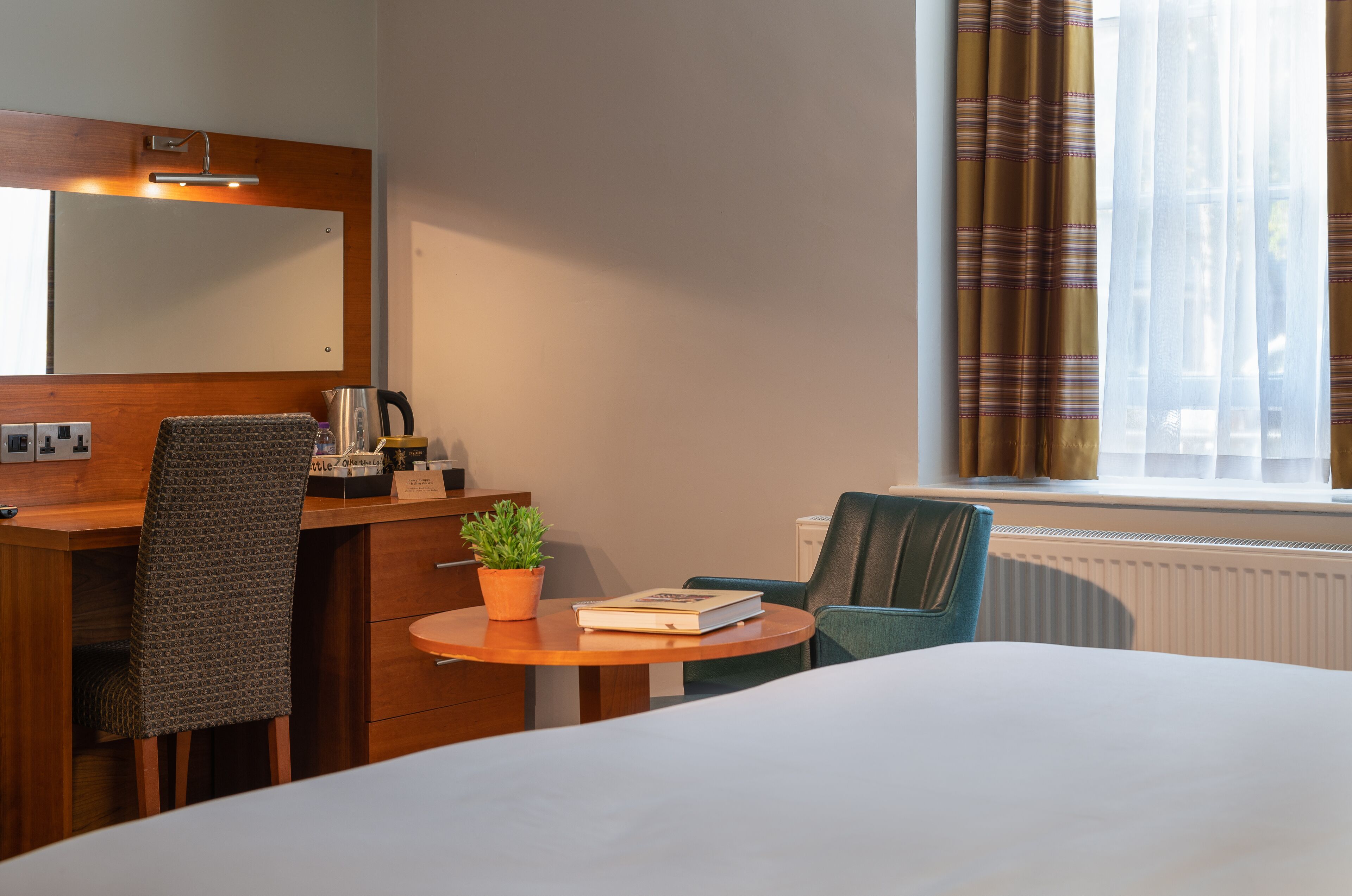 A hotel room with a bed, a desk with a chair, a small table with a plant, a book, and a coffee maker.