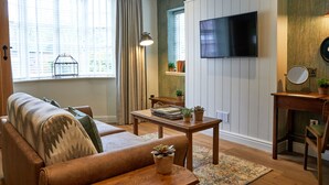 A living room with a sofa, a coffee table, a TV mounted on the wall, a window with curtains, and a small table with plants.