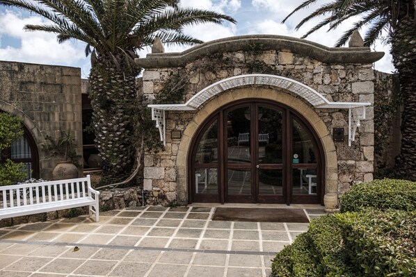 A stone building with a large arched entrance, a white bench, and a palm tree. - Hotel Ta' Cenc & Spa (Sannat)
