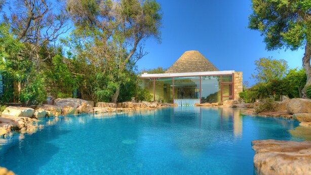Una piscina azul cristalina rodeada de rocas y árboles, con un edificio moderno de piedra al fondo.