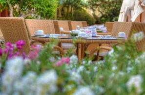 A dining table set with white tableware, surrounded by wicker chairs, in a garden setting with flowers. - Hotel Ta' Cenc & Spa (Sannat)