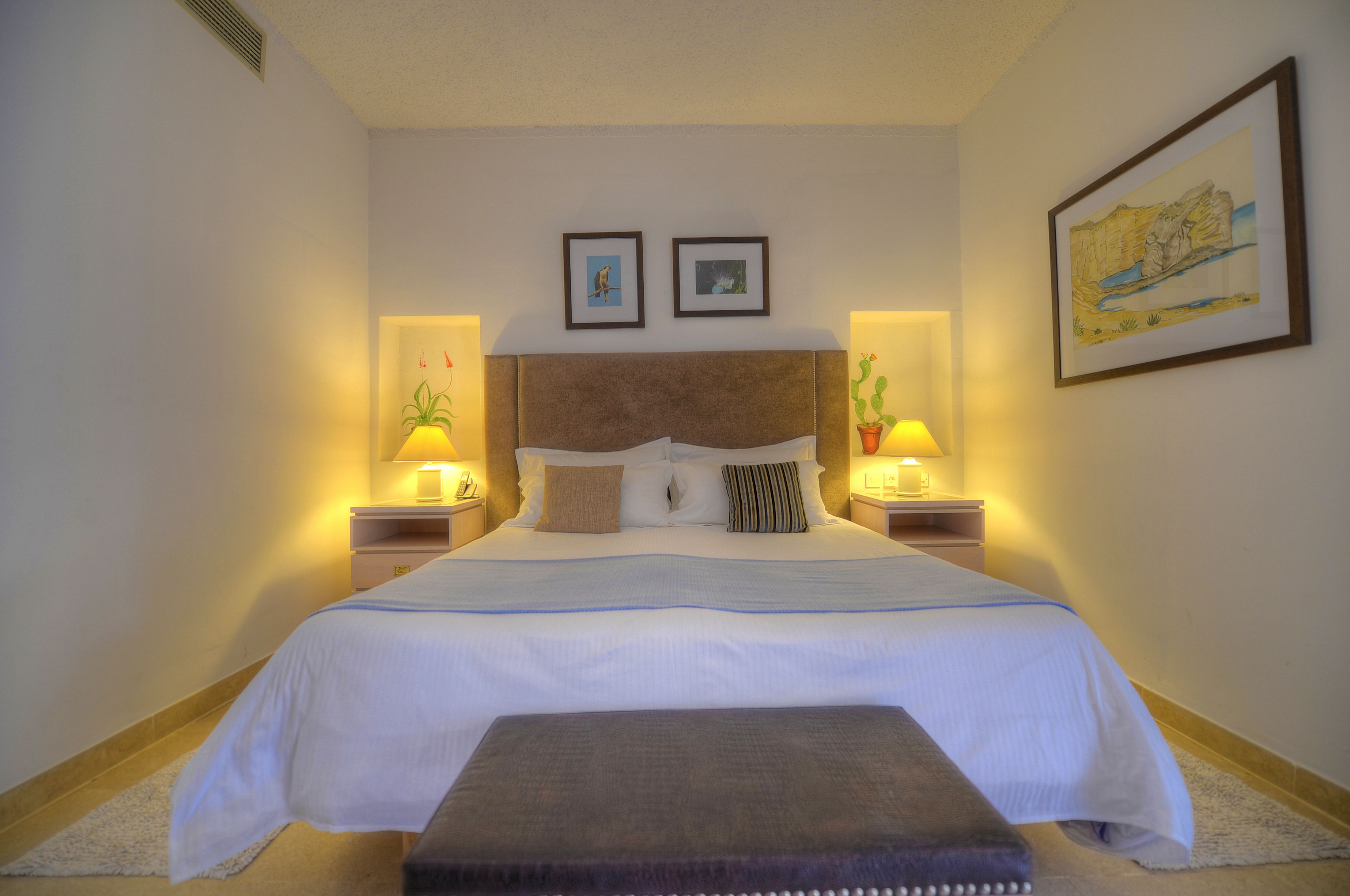 Uma cama bem arrumada com cabeceira marrom, duas mesas de cabeceira com luminárias e um quadro emoldurado sobre a cama.