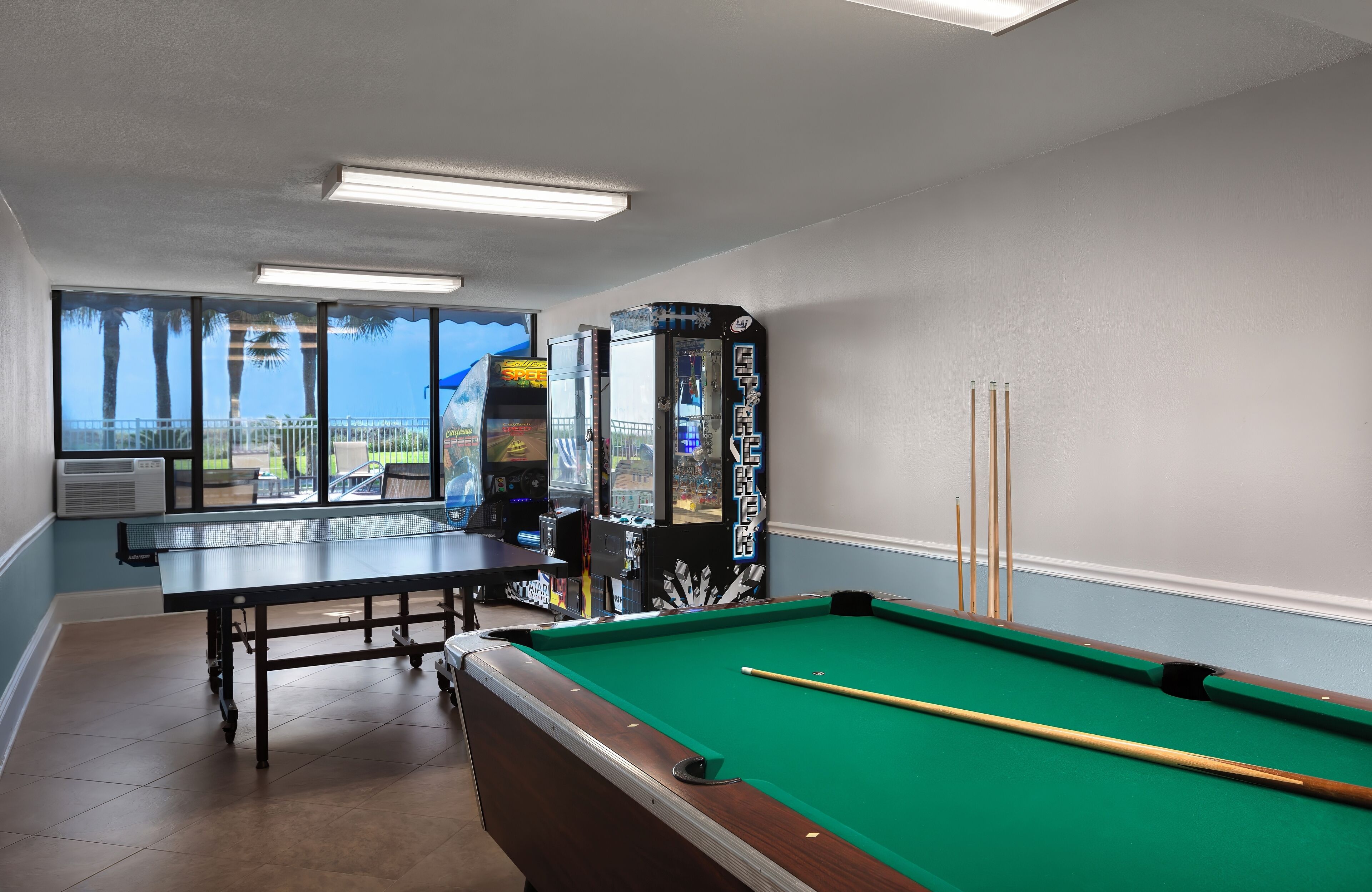 Een pooltafel met een groene viltbekleding, een poolkeu, en een tafeltennistafel in een ruimte met grote ramen en uitzicht op palmbomen.