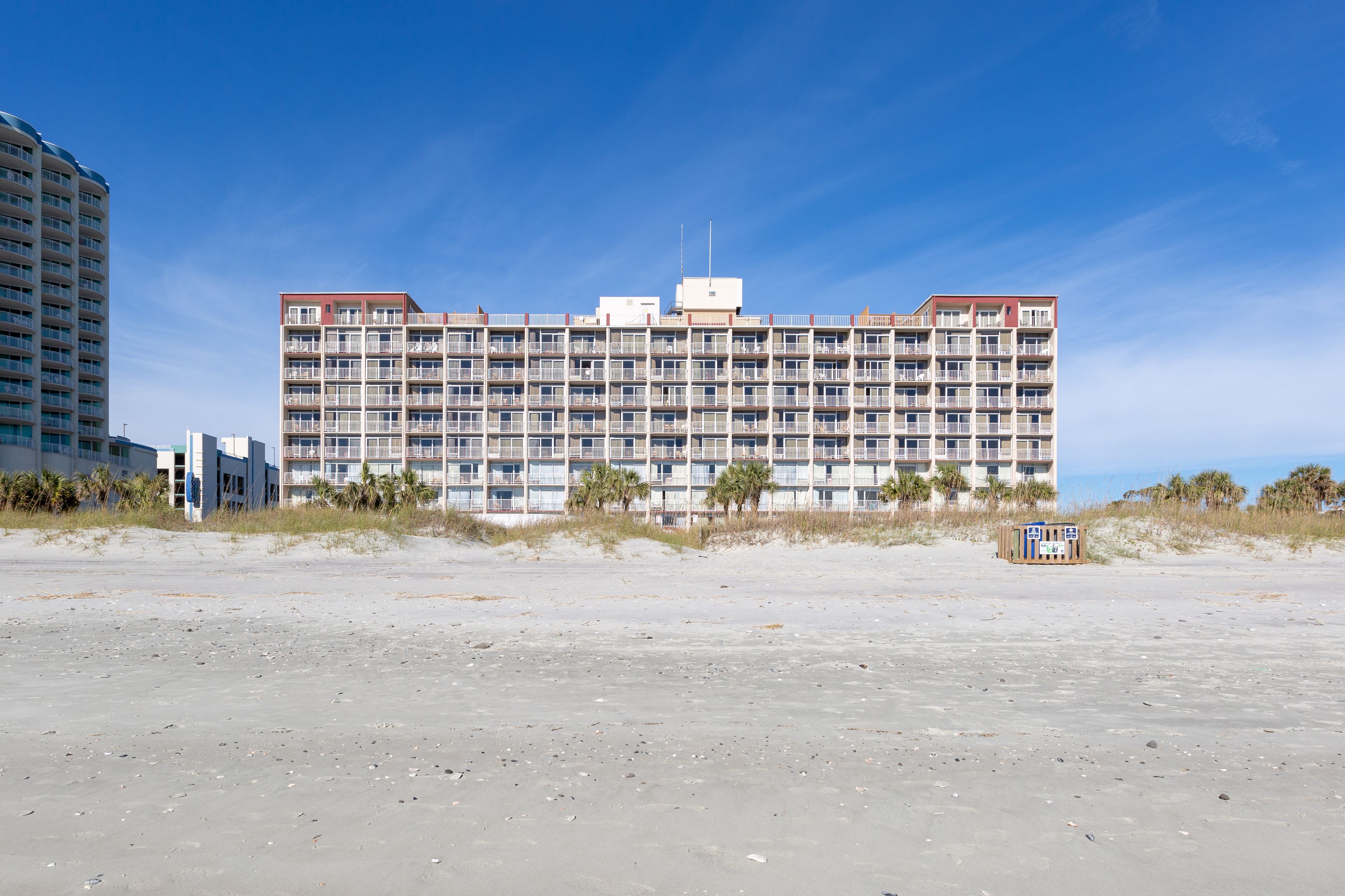 A large, multi-story hotel with a flat roof and numerous balconies, situated on a sandy beach with palm trees in the foreground.