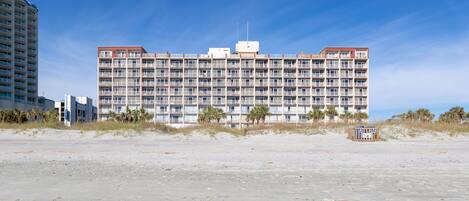 A large, multi-story hotel with a flat roof and numerous balconies, situated on a sandy beach with palm trees in the foreground.