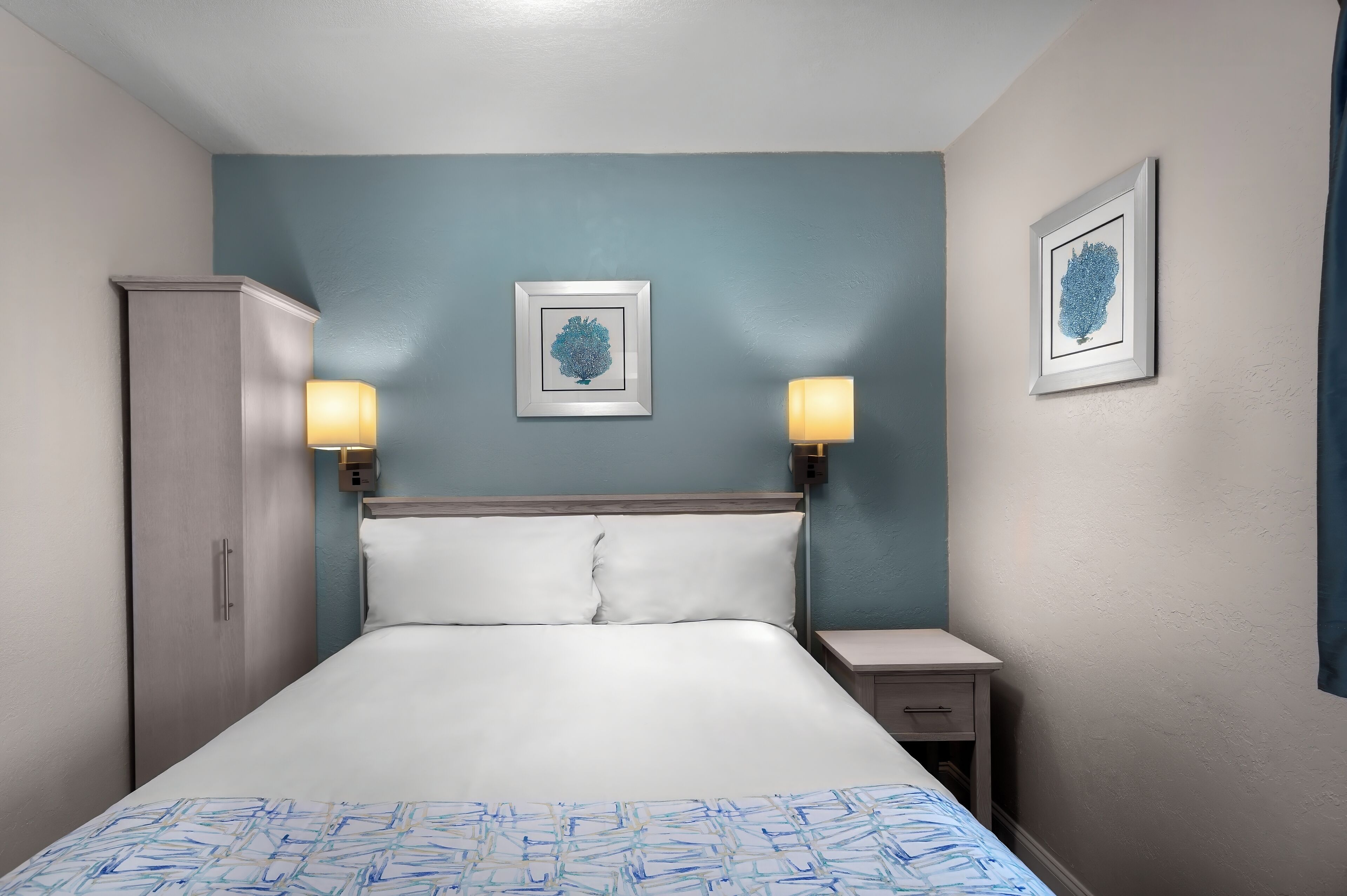 A neatly made bed with white linens, two bedside lamps, and two framed artworks on the wall.