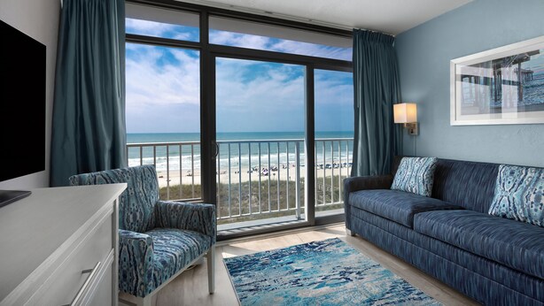 Ein Zimmer mit einer großen Schiebeglastür, die Blick auf einen Strand bietet, eine Couch, ein Sessel und ein Flachbildfernseher.