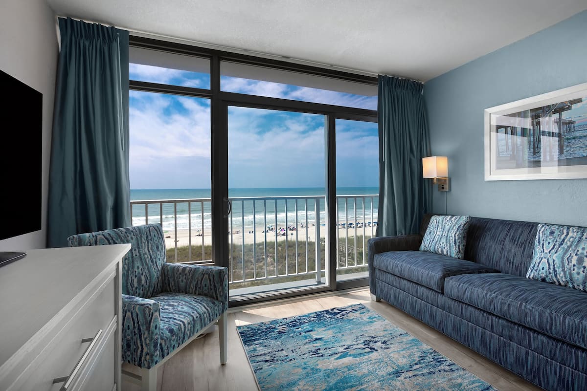 A room with a large sliding glass door leading to a beach view, a sofa, a chair, and a flat-screen TV.