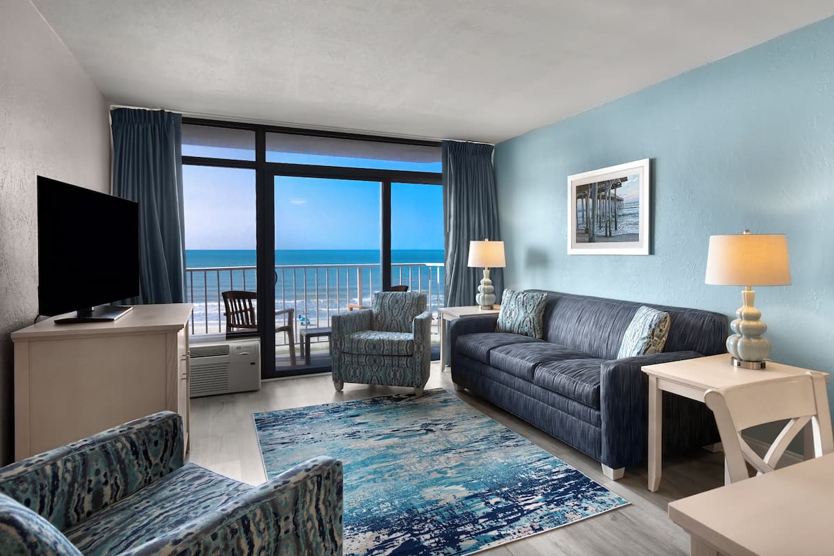 A hotel room with a balcony, a TV, a sofa, two armchairs, a coffee table, and a rug.