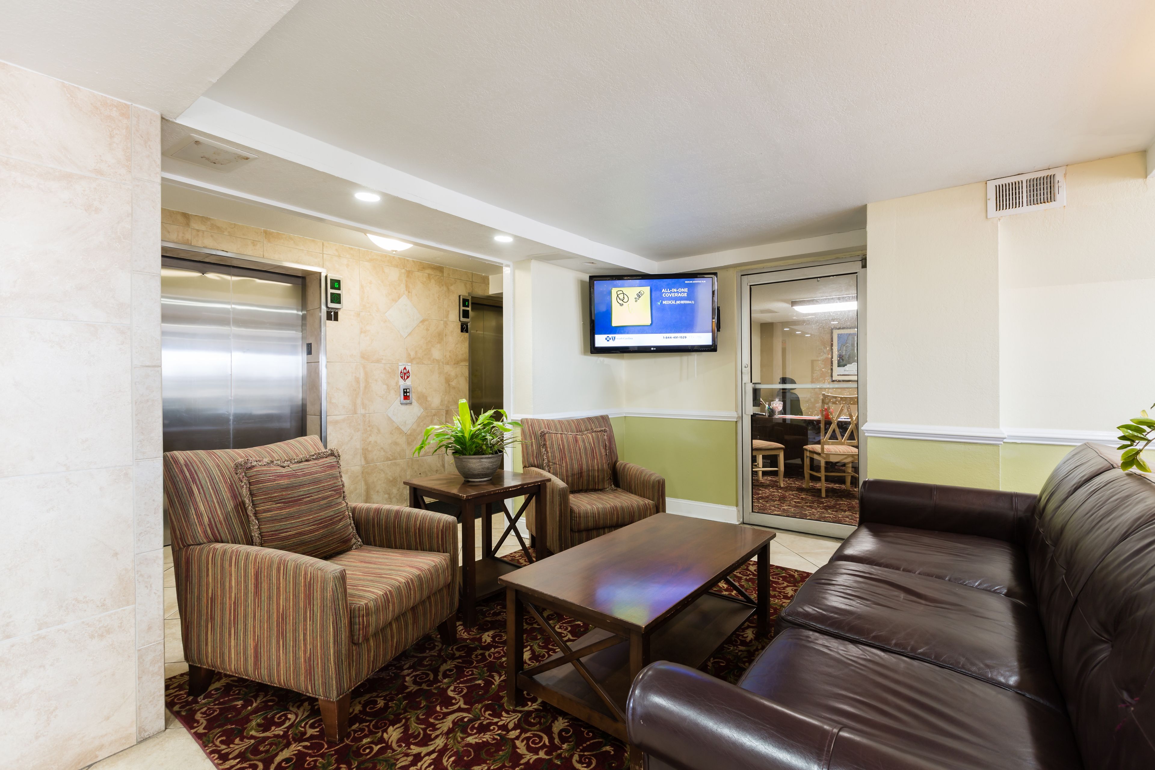 A lobby with a brown leather sofa, striped armchairs, a wooden coffee table, and a potted plant. An elevator is visible in the background.