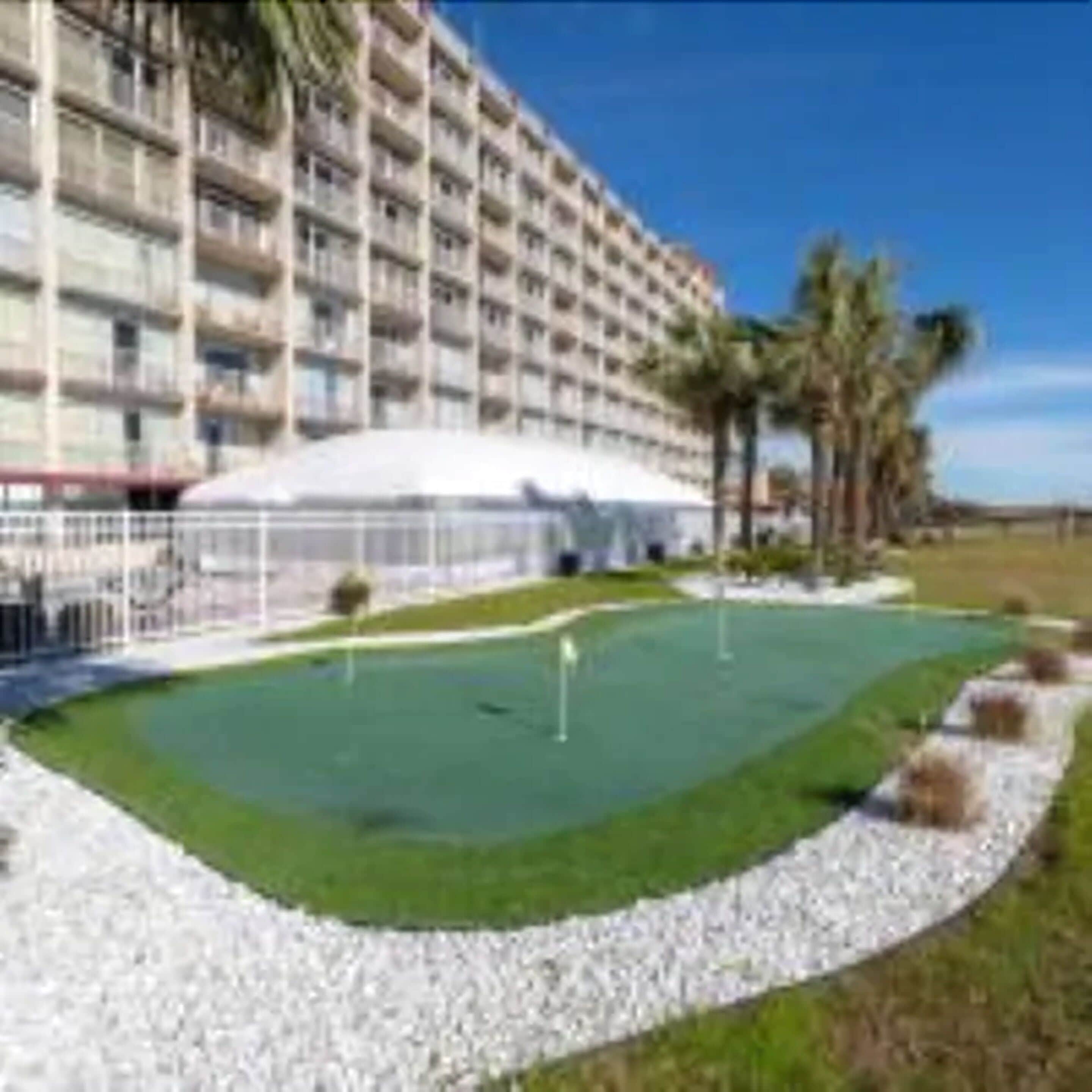 A hotel with multiple floors, a pool area with a green lawn, and palm trees.