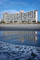 Un hotel frente a la playa con varios balcones, una playa arenosa en primer plano y un cielo despejado.