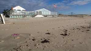 Une maison de plage avec une façade en verre, un drapeau et une plage de sable où l’on peut voir des empreintes de pas.
