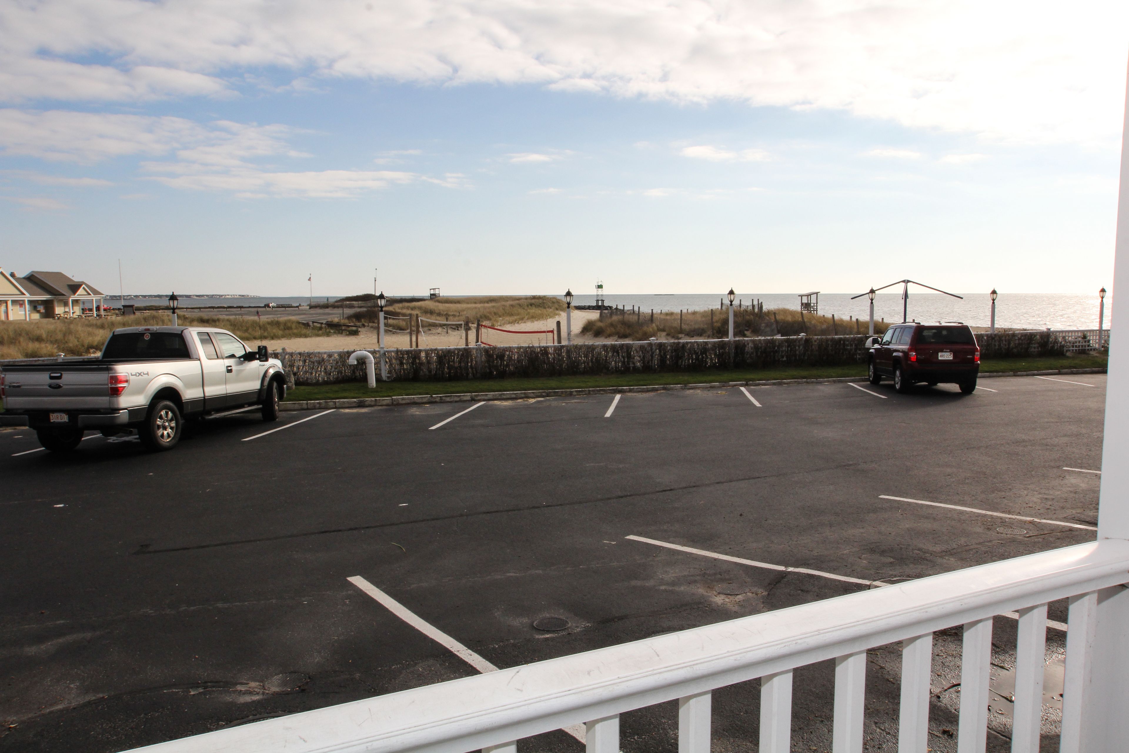 A parking lot with two vehicles, a white pickup truck and a red SUV, near a beach with a stone wall and a fence.