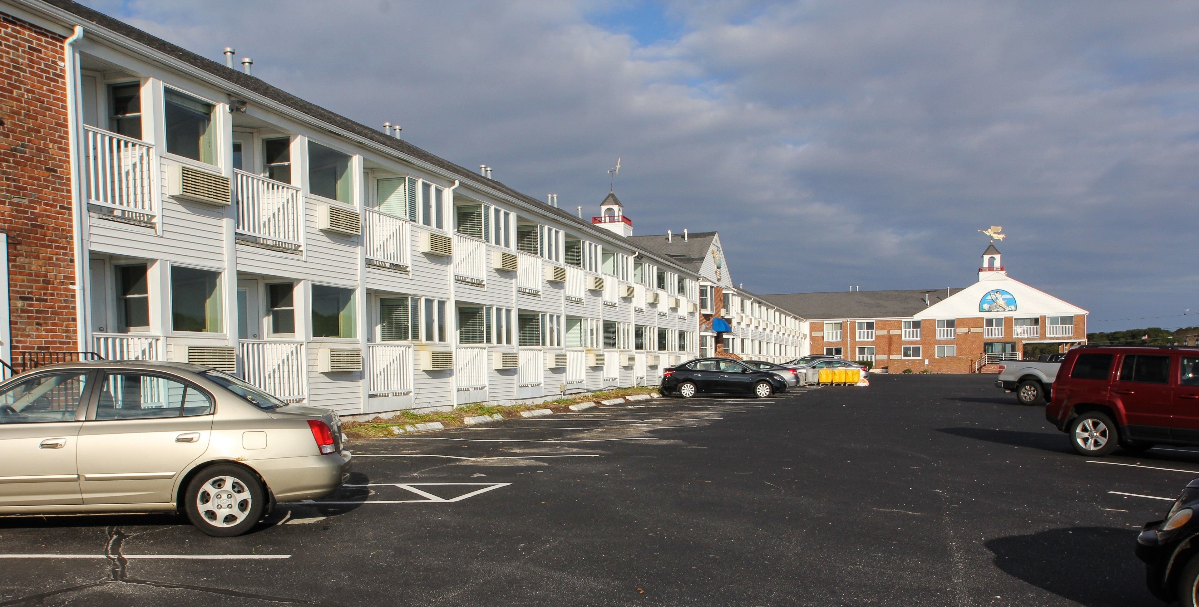 A parking lot with several cars, a building with multiple windows and balconies, and a church with a steeple in the background.