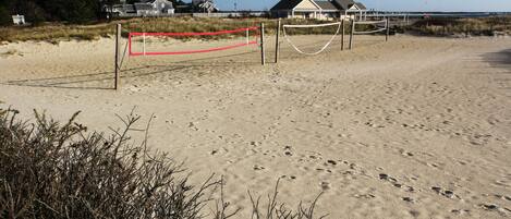 Una spiaggia con un campo da pallavolo con rete rossa, circondato da sabbia e vegetazione rada.