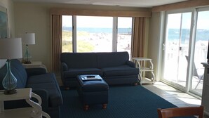 Un salon comprenant un canapé, un pouf et une table à manger avec des chaises. Une grande fenêtre offre une vue sur la plage et l’océan.