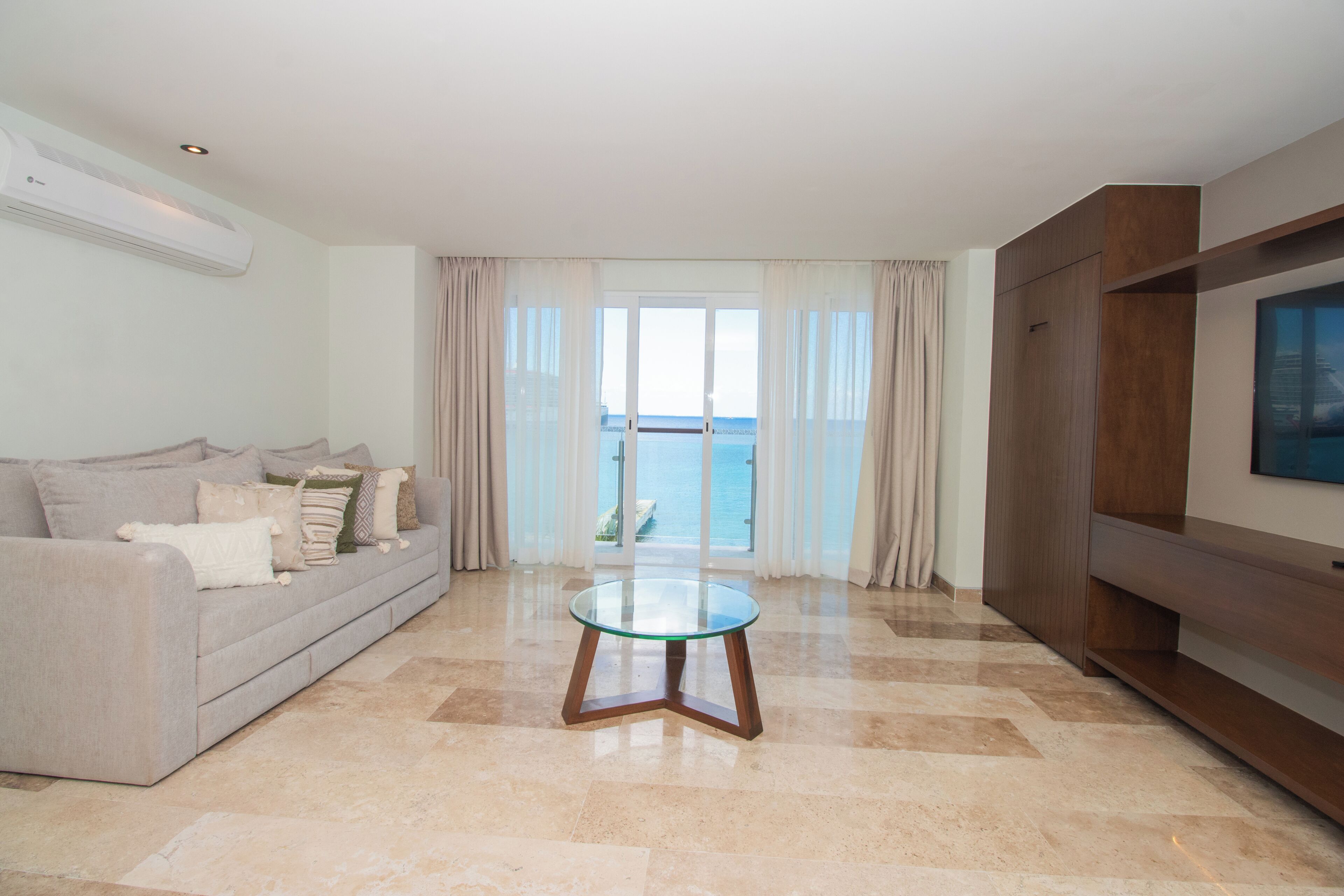 Un salon comprenant un canapé, une table basse avec un plateau en verre, un téléviseur à écran plat et une vue sur la mer par une porte vitrée.