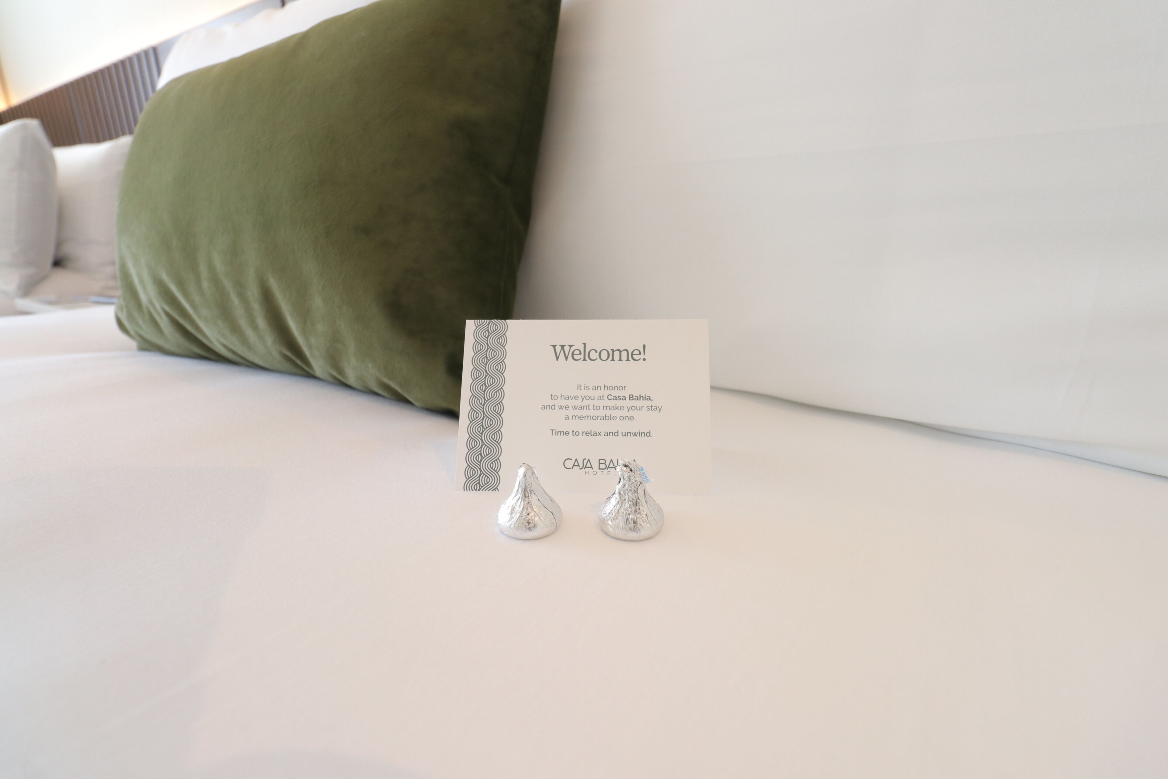 A bed with a white pillow, a green pillow, a welcome card, and two silver chocolates.