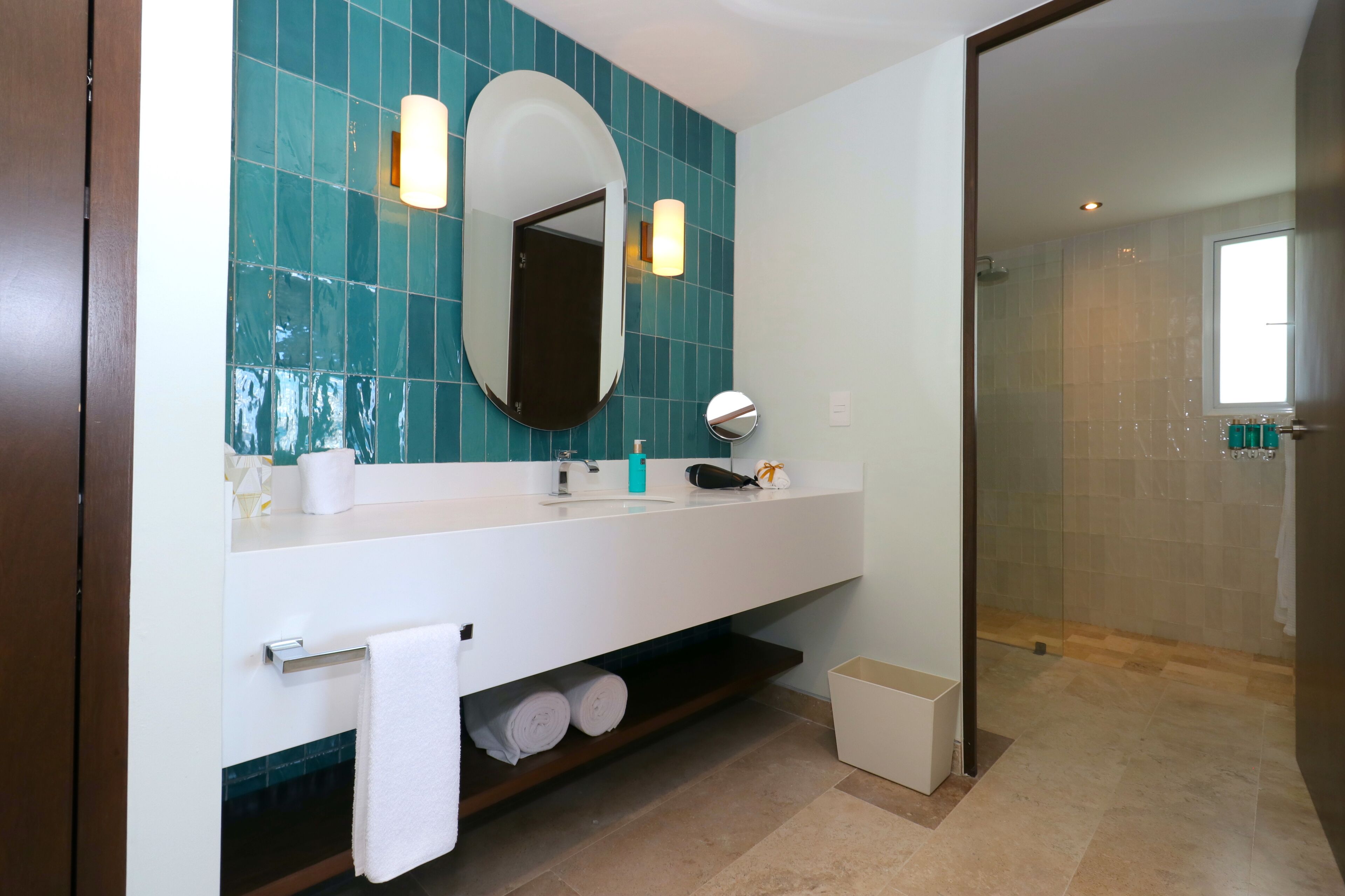 Un baño moderno con un espejo grande, tocador blanco, azulejos verde azulado, cabina de ducha de vidrio y un pequeño cubo de basura.