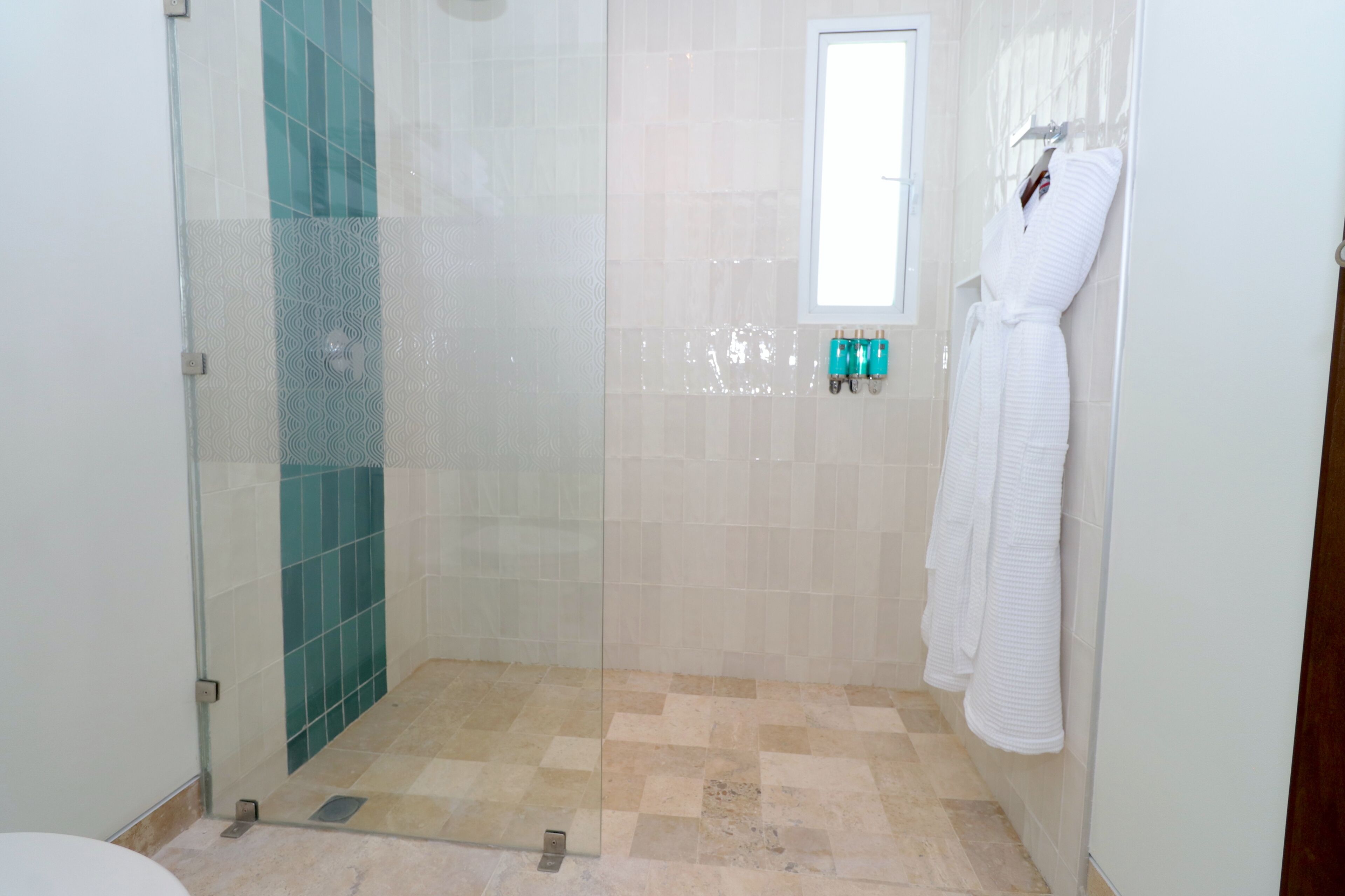 Un baño con ducha envuelta en vidrio, paredes con azulejos, una ventana y un perchero con toallas blancas.