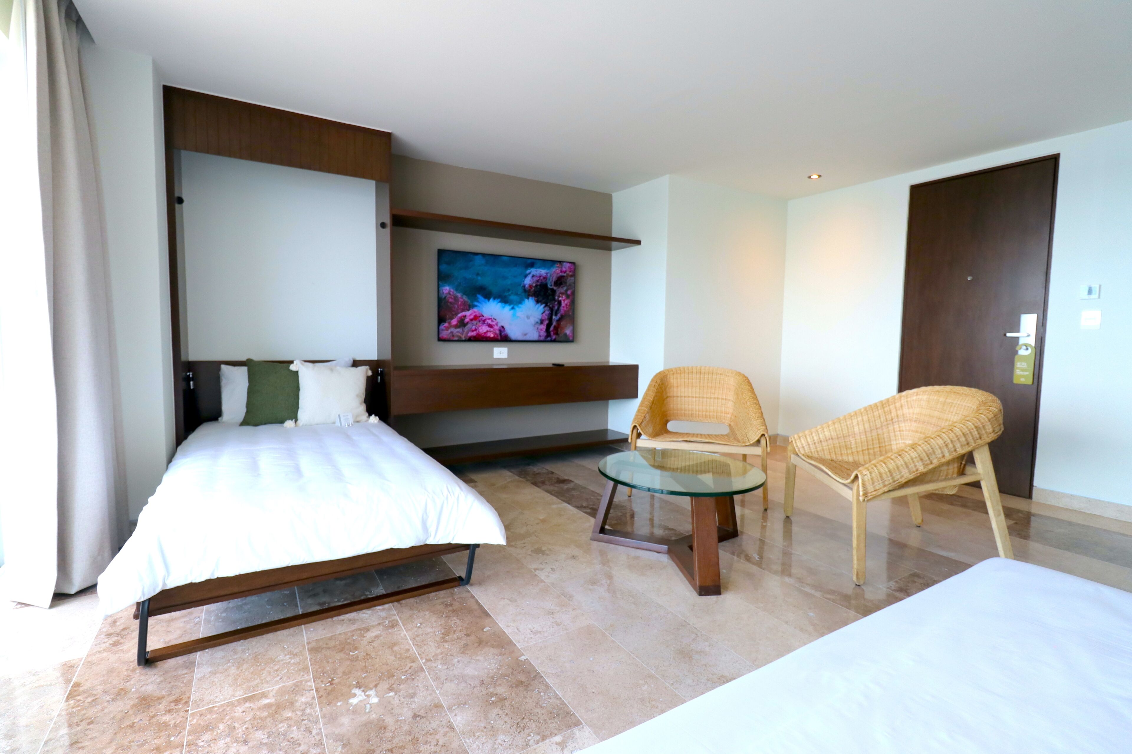 Een moderne hotelkamer met een bed, een televisie, twee rieten stoelen en een glazen tafeltje.