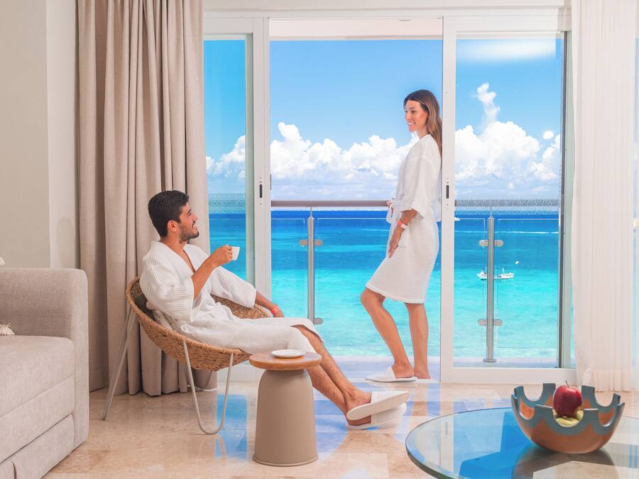 A room with a sofa, a wicker chair, a glass table, and a view of the ocean through large windows.
