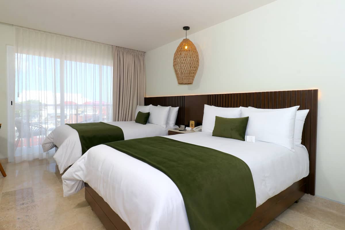 Habitación de hotel con dos camas, un ventanal con cortinas, una lámpara colgante y vista a la ciudad.