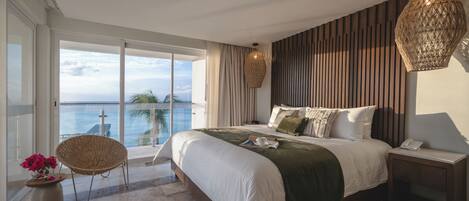 A bedroom with a large bed, a nightstand, a wicker chair, and a view of the sea.