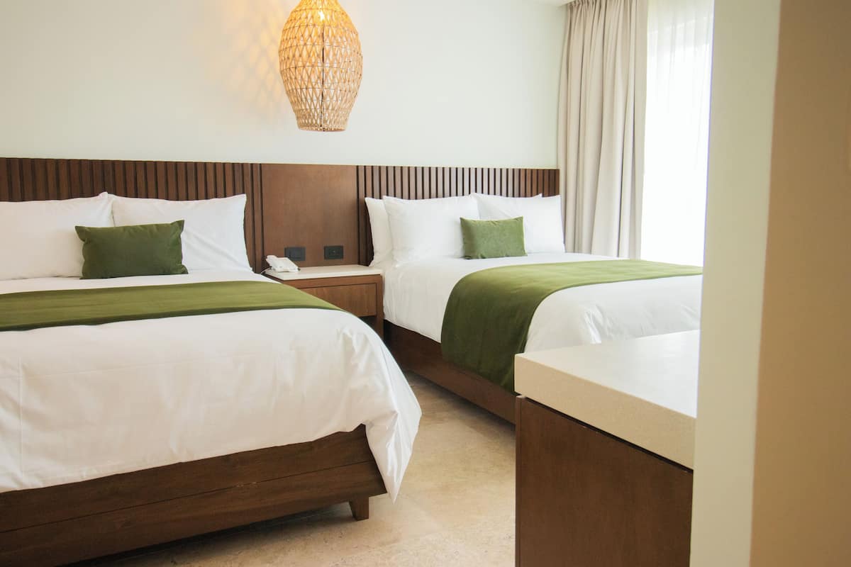 Habitación de hotel con dos camas, una mesita de noche, una lámpara y un ventanal con cortinas.