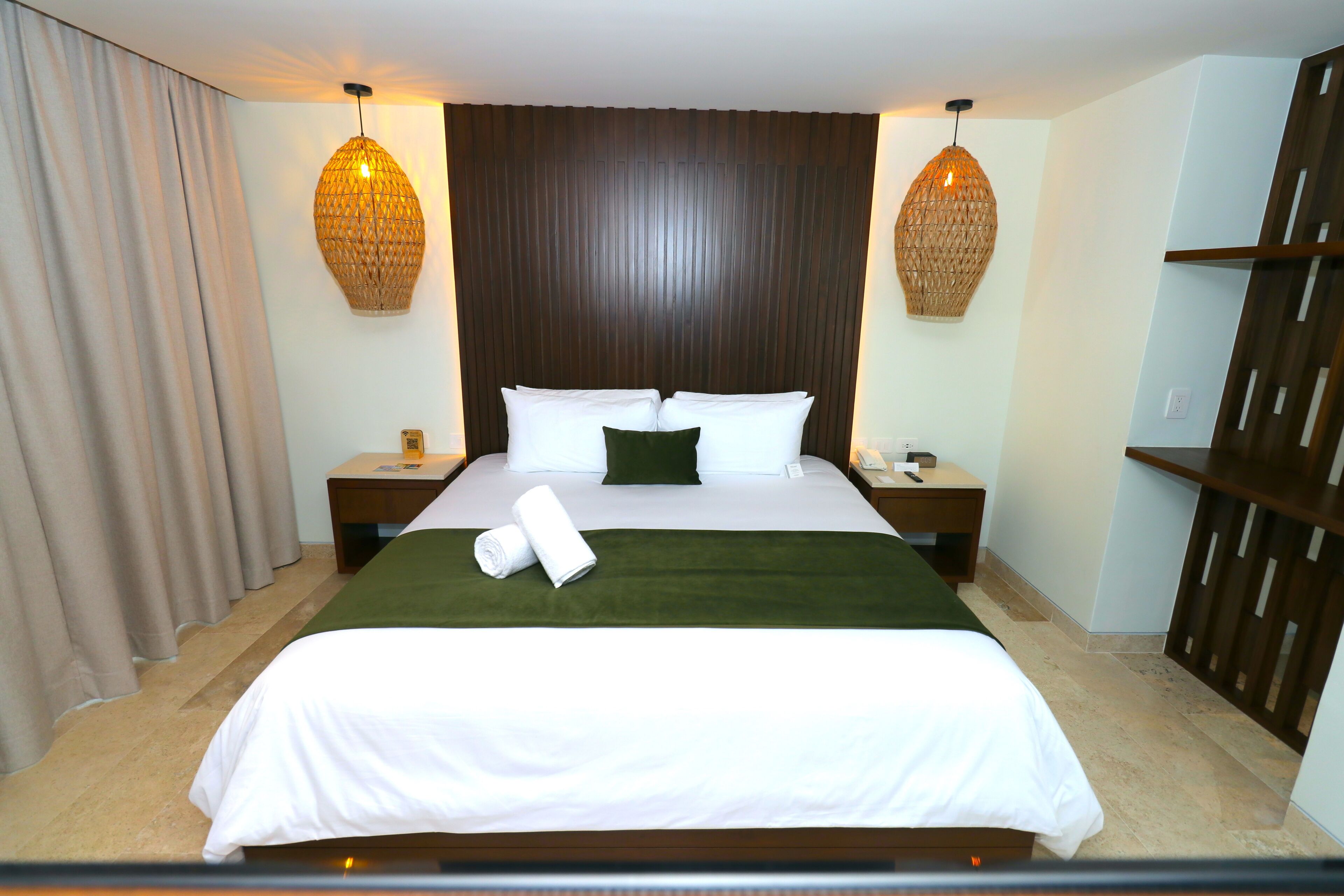 A hotel room with a large bed, two bedside tables, a green throw blanket, and two hanging lamps.