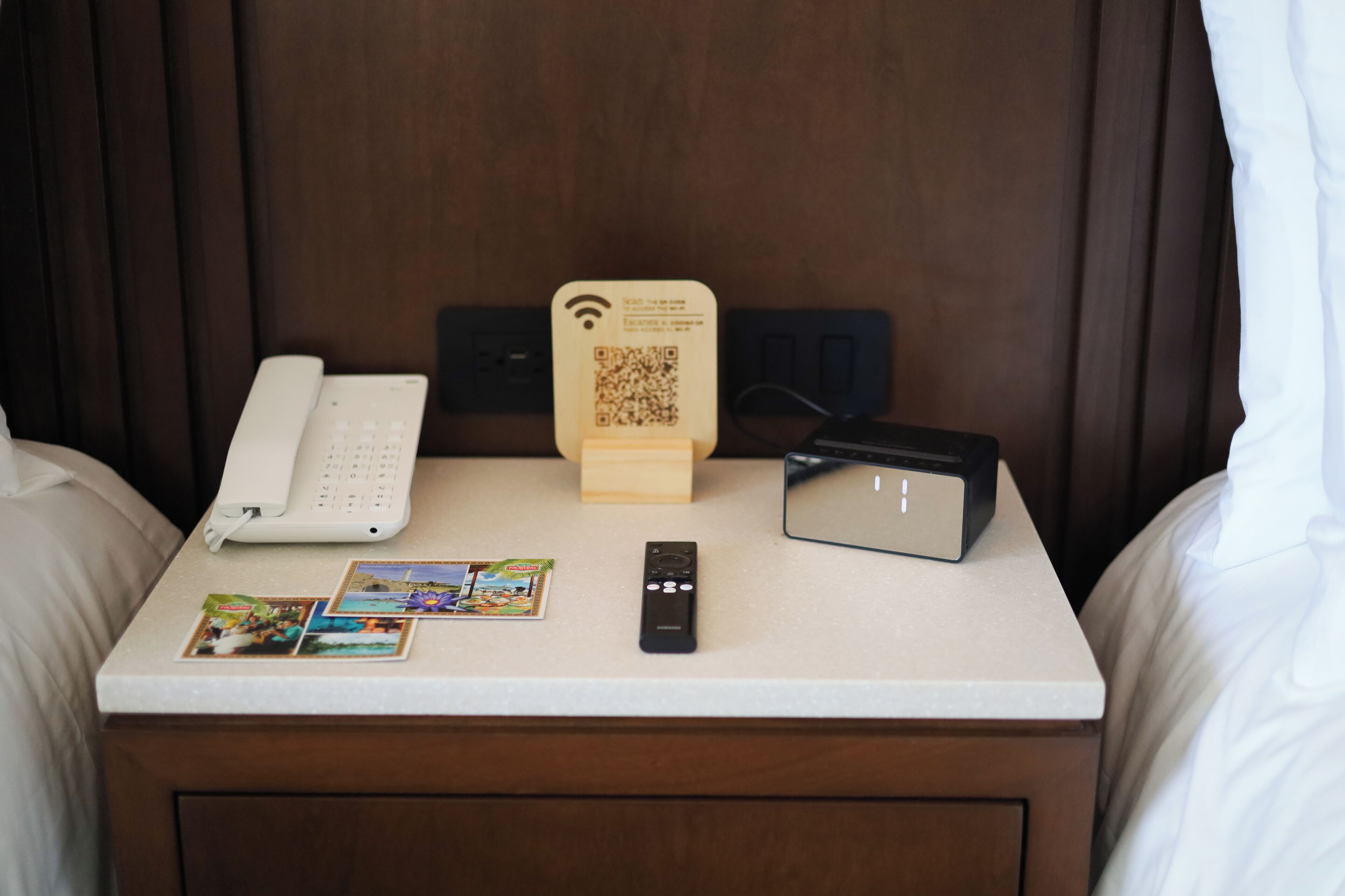 A hotel room with a bedside table featuring a white phone, a remote control, a QR code stand, and a small booklet.