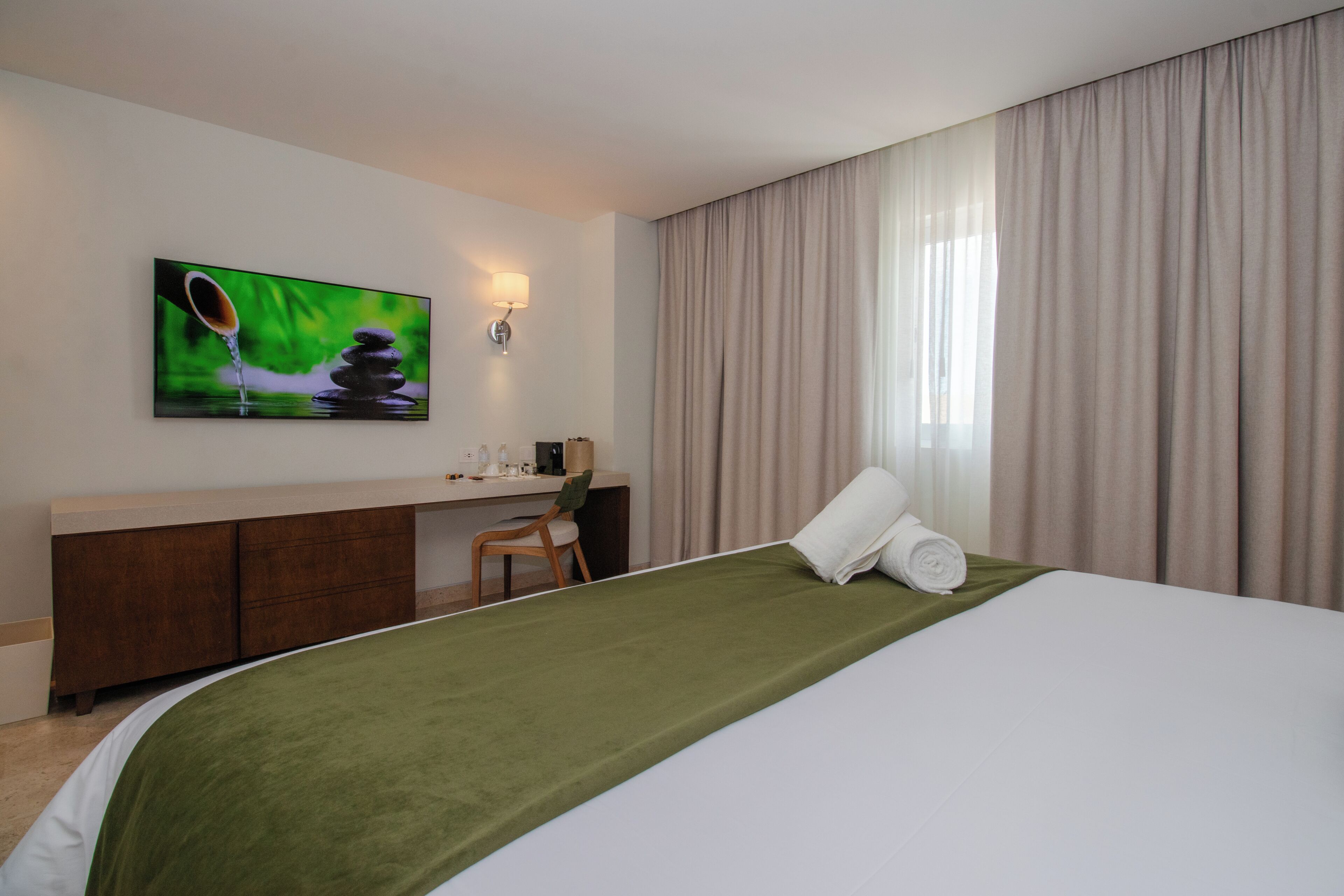 A hotel room with a bed, a desk, a chair, a painting, and a window with curtains.