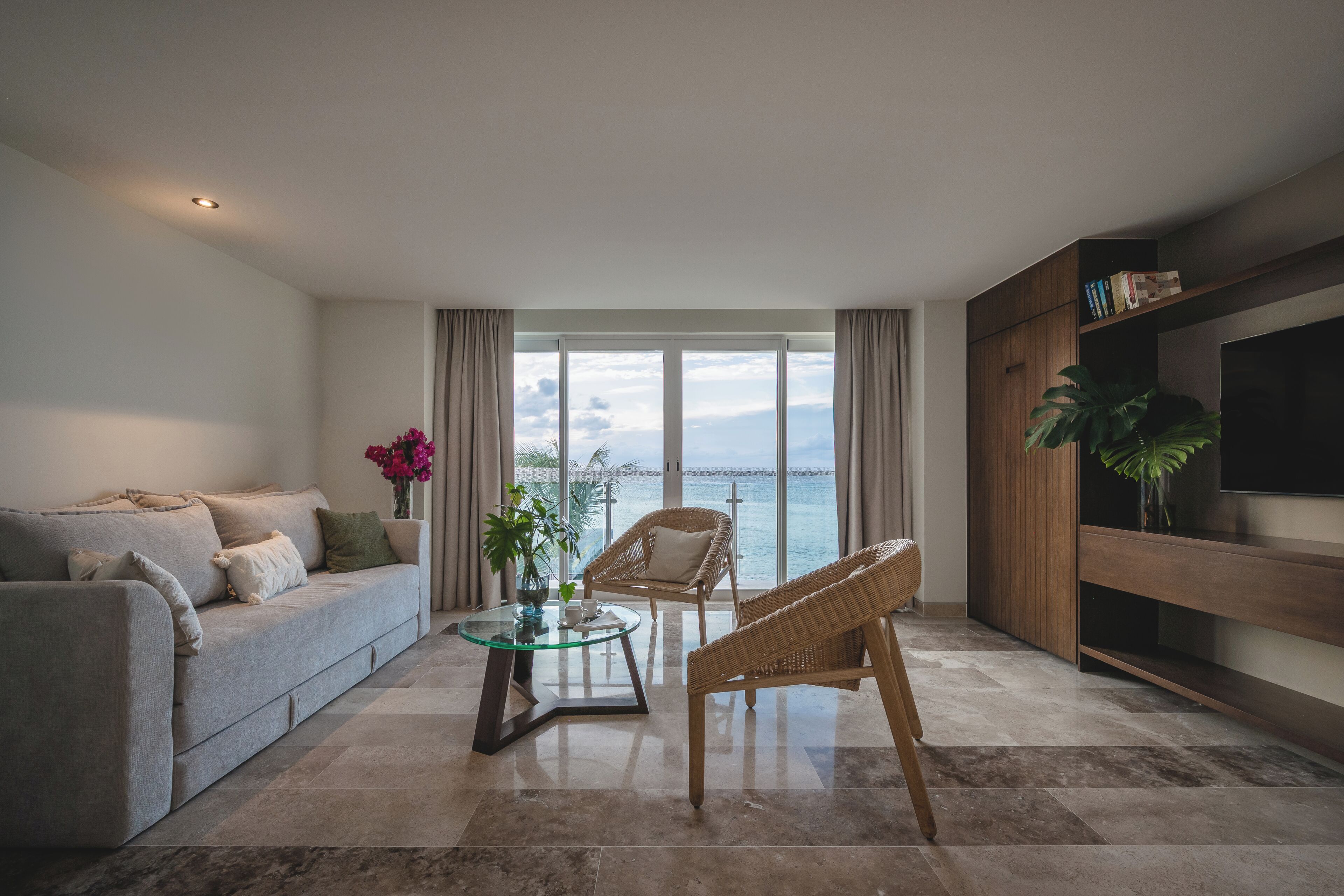 Een moderne woonkamer met een bank, een glazen salontafel, rieten stoelen en een televisie op een houten dressoir.