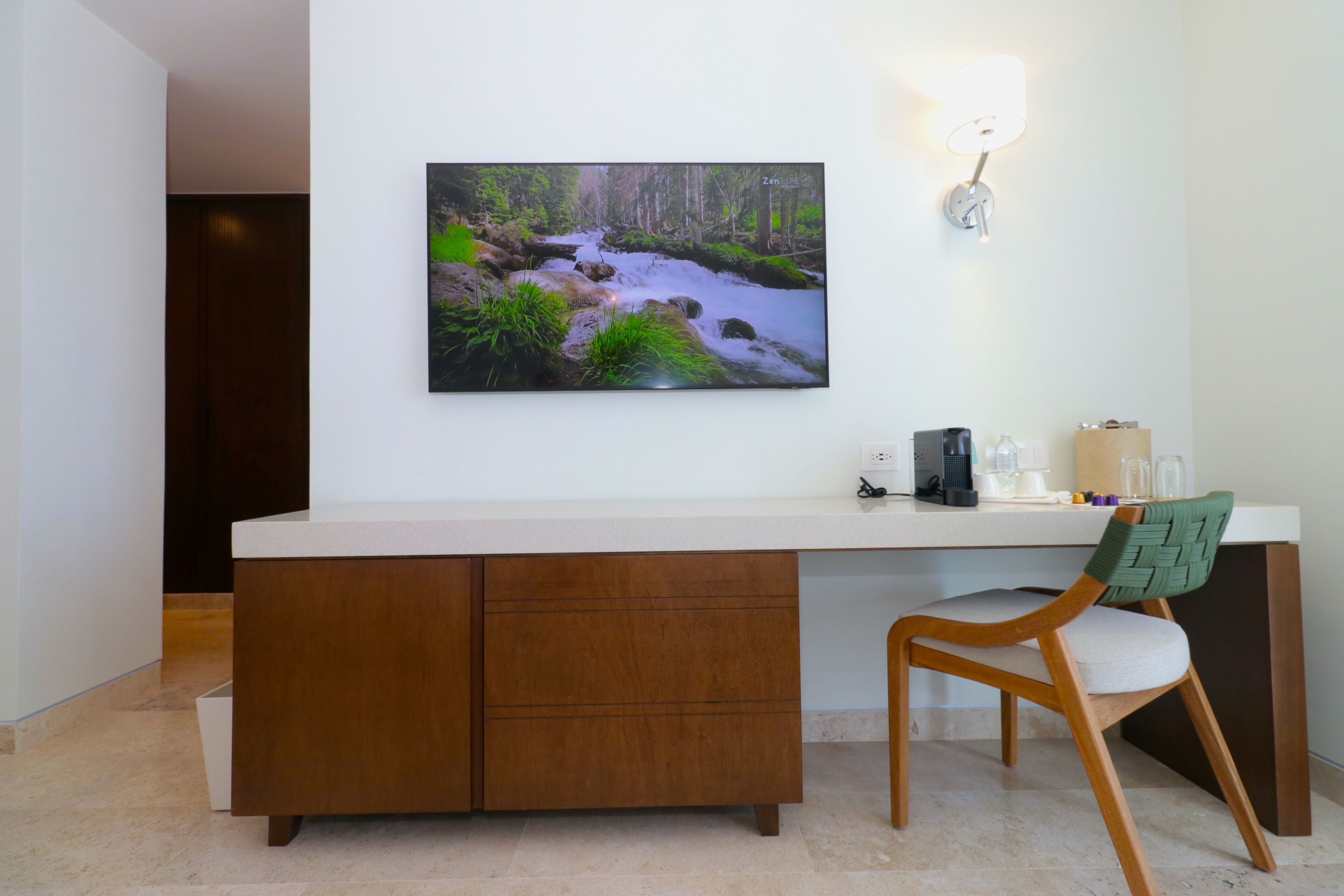 Una habitación moderna con un escritorio de madera, una silla, un cuadro colgado en la pared que muestra un arroyo en el bosque, y una lámpara de pared.