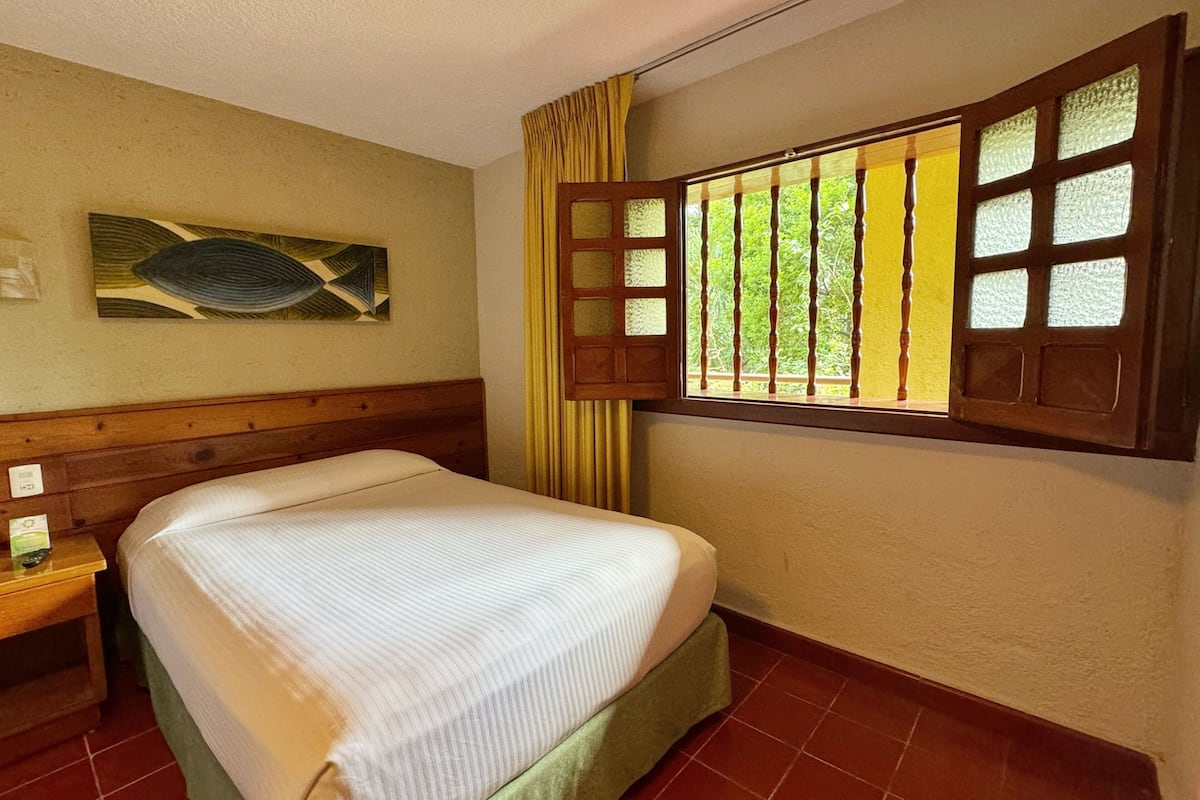 Een hotelkamer met een bed, een nachtkastje, een raam met houten luiken en een schilderij aan de muur.