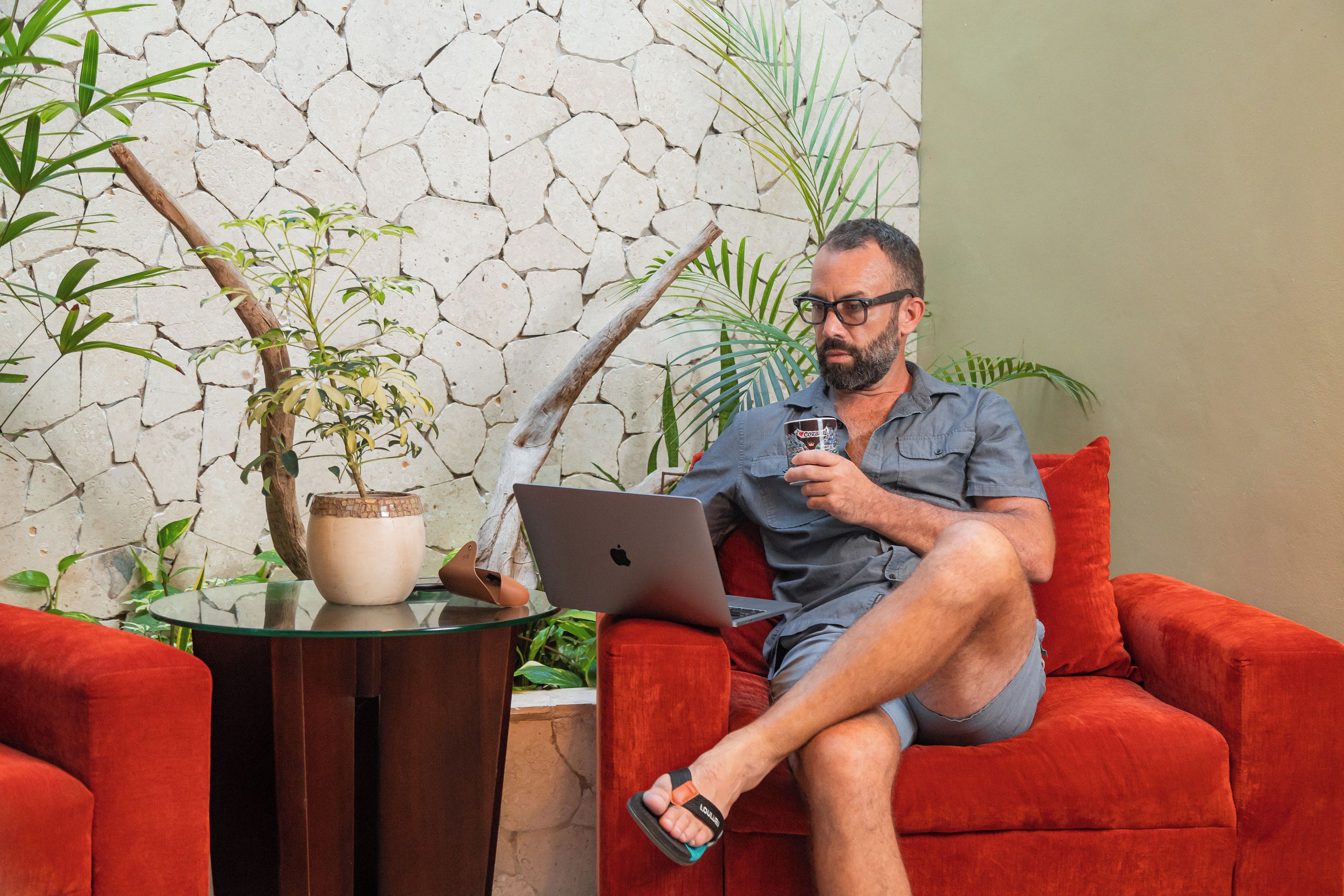 A man sitting on a red couch with a laptop, holding a mug, and surrounded by plants and a stone wall.