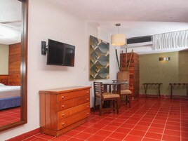 A hotel room with a wooden dresser, a TV, a dining table with chairs, a red tiled floor, and a decorative wall piece.