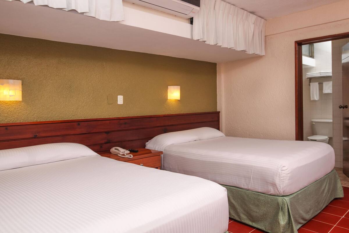 Habitación de hotel con dos camas, cabecero de madera, mesita de noche con teléfono y baño visible a través de una puerta abierta.