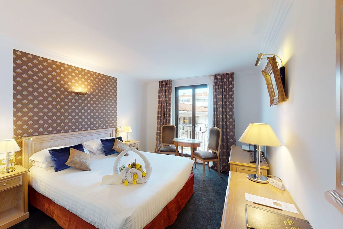 A hotel room with a large bed, two bedside tables, a desk, a chair, and a window with curtains.