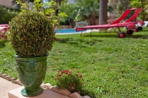A well-maintained garden with a neatly trimmed bush in a green pot, a small flowering shrub, and a red lawn chair in the background. - Sun Riviera (Cannes)